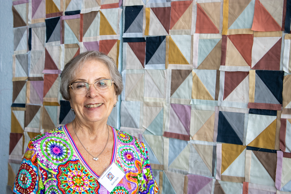 Quilt retreat participant in colorful embroidered V-neck top, standing before patchwork quilt
